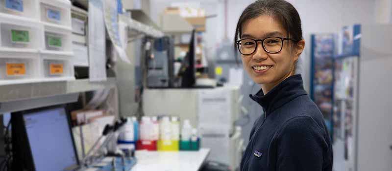 Mei San Tang smiles at the camera while sitting in front of a computer in a lab.
